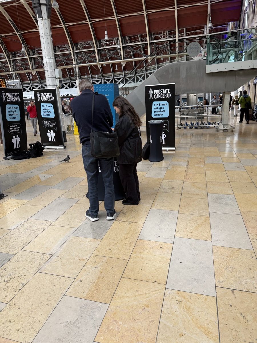 CKT brand ambassador working a station shift, branded Prostate Cancer UK stands behind.