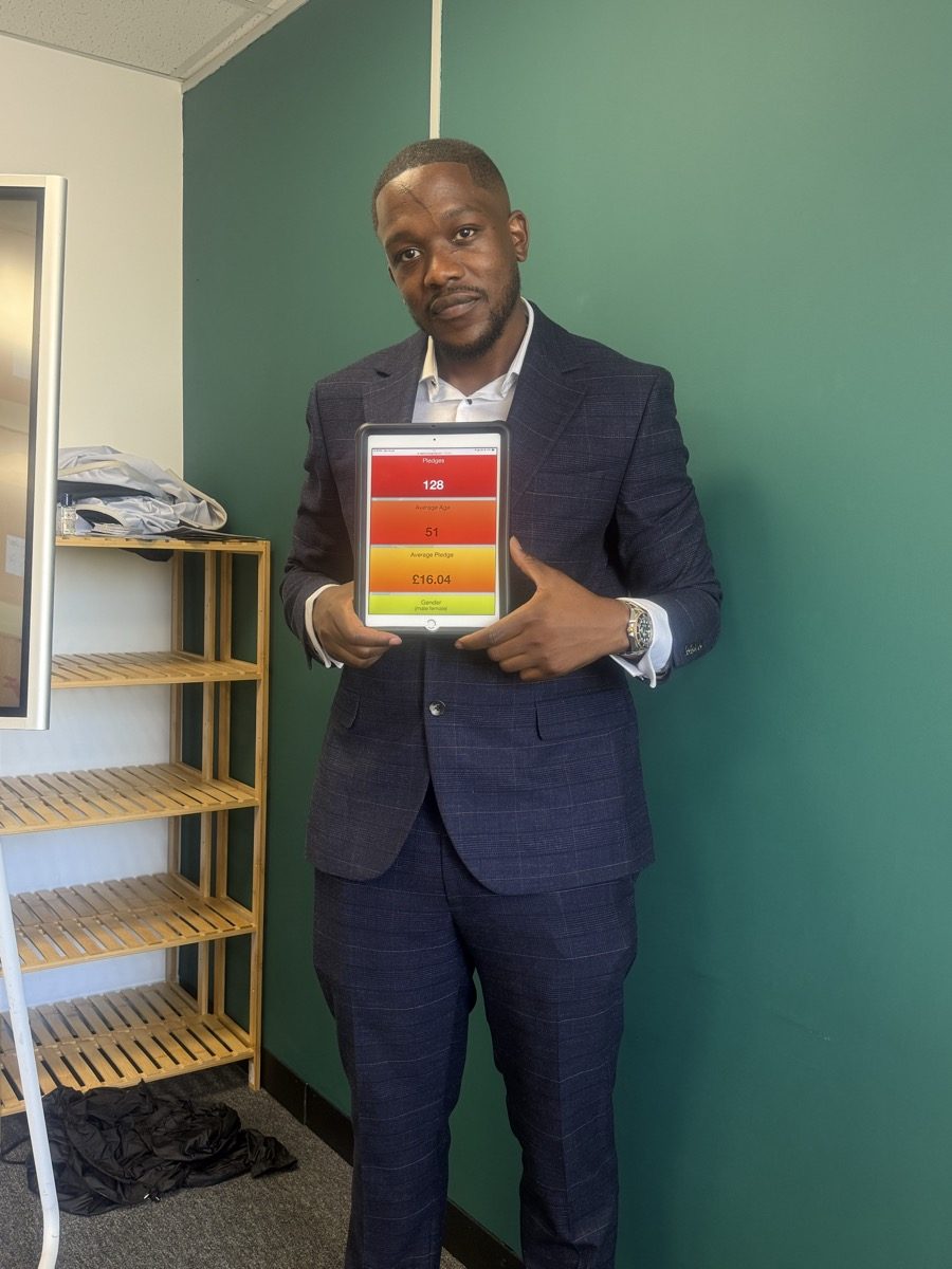 Tunde Balogun holding a tablet showing 128 pledges in one week — an average pledge of £16.04 per donor — recorded at Waterloo Station.
